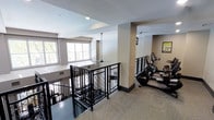 This fitness center is a thoughtfully designed exercise space that combines functionality with a clean, modern aesthetic. The layout is open and airy, benefiting from large windows that allow generous amounts of natural light to fill the room, creating a bright and inviting atmosphere for workouts. The space is anchored by a sturdy staircase enclosed with distinctive black metal diamond-patterned railings, providing access to the level below and serving as a visual focal point within the space. A structural column helps define the exercise zone, where a row of cardio equipment — including elliptical machines and steppers — is neatly arranged along the wall. Framed artwork adds a subtle decorative touch to the neutral-toned walls, which are painted in a warm beige palette that complements the speckled carpet flooring throughout the fitness area. Recessed and pendant lighting supplements the natural light, ensuring the space remains well-lit at all times. The overall ambiance is practical yet polished, offering users a comfortable and well-maintained environment suited for regular physical activity. The open plan and considered layout make this fitness center an accessible and efficient space for a range of exercise routines.
