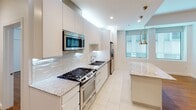 This well-designed single-floor residence offers a clean, modern aesthetic throughout, with thoughtful finishes and a consistent design language across every room. The kitchen is a standout feature, showcasing white flat-panel cabinetry that extends to the ceiling, granite countertops, a textured subway tile backsplash, and a full suite of stainless steel appliances including a gas range, built-in microwave, and refrigerator. A kitchen island provides additional counter space, while pendant lighting adds a warm, stylish touch above the adjacent dining area. The open living area flows naturally from the kitchen and is bathed in natural light from large windows. Rich hardwood flooring extends throughout the living spaces and bedrooms, lending warmth and cohesion to the interior. The bedrooms are generously sized, each featuring tray ceilings, ample wall space, and wall-mounted split-system air conditioning units for individual climate control. Two bathrooms serve the residence, both finished with large-format white floor tile, frameless sliding glass shower enclosures with textured tile surrounds, dark-toned vanities with marble-look countertops, and framed mirrors. One bathroom features a combined tub and shower, while the other offers a standalone walk-in shower. The overall ambiance is clean, contemporary, and move-in ready.
