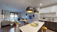 This thoughtfully designed single-floor residence showcases a seamless open-concept layout that connects the kitchen, dining, and living areas into one cohesive, light-filled space. The gourmet kitchen is a standout feature, boasting two-tone cabinetry with high-gloss white uppers and rich wood-grain lowers, quartz countertops, stainless steel appliances, gold hardware accents, and a central island with bar seating. Striking oversized globe pendant lights in brushed metal anchor the kitchen and dining zone with a bold, contemporary flair. The living room offers a relaxed, sophisticated atmosphere with large sliding glass doors opening to a private balcony, inviting abundant natural light and outdoor views. The primary bedroom is a serene retreat, also featuring direct balcony access through sliding glass doors. Its en-suite bathroom includes a walk-in glass shower, quartz vanity, and wood-tone cabinetry. A second bedroom benefits from generous natural light through large windows. The secondary bathroom features tiled walls, a soaking tub with shower, and an adjacent closet with built-in shelving. Rich hardwood-style flooring flows throughout the main living spaces, while neutral tones and modern finishes create a cohesive, contemporary ambiance across the entire residence.