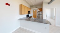 This single-floor urban unit offers a well-organized layout that balances open living with functional design. Light hardwood-style flooring flows throughout, creating a cohesive and airy feel across all areas.

The kitchen is a standout feature, designed in an open-concept style with a large granite-topped peninsula that doubles as a breakfast bar. Warm wood cabinetry lines the walls, providing ample storage, while stainless steel appliances — including a range and refrigerator — along with a built-in microwave complete the setup. Pendant lighting hangs above the peninsula, and exposed piping along the ceiling adds an industrial loft-like character.

The living area benefits from generous natural light, with large black-framed windows that frame urban rooftop views of the surrounding cityscape. The high ceilings and open sightlines enhance the sense of space, giving the area a bright and expansive feel.

The bathroom is clean and well-appointed, featuring a tub and shower combination enclosed by sliding frosted glass doors, a dark-cabineted vanity with a white sink, a large mirror with lighted fixtures above, and light tile-style flooring that coordinates with the rest of the unit.

Overall, this space presents a modern urban aesthetic with industrial accents, practical finishes, and an abundance of natural light.
