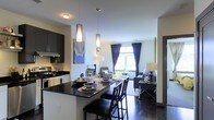 This thoughtfully designed single-floor residence showcases a seamless open-concept layout that connects the kitchen, dining, and living areas into one cohesive, flowing space. The modern kitchen is outfitted with dark cabinetry, a sleek gas range with hood vent, and a dark granite-topped breakfast bar that doubles as a dining counter. Stylish cylindrical pendant lights hang above the bar, adding a contemporary focal point to the space while providing warm, targeted illumination. The dining area is furnished with high-back bar seating and a well-appointed table set, creating an inviting atmosphere for entertaining. Beyond the kitchen and dining space, the living room is generously proportioned with room for multiple seating arrangements. A large window dressed with deep navy curtains allows natural light to pour in, brightening the light-colored walls and dark flooring throughout. The overall aesthetic is modern and sophisticated, with a carefully curated palette of dark tones balanced by warm lighting and neutral walls. The open floor plan promotes a sense of spaciousness and connectivity, making this residence both functional and visually striking.