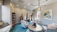 Located at 7009 Almeda Rd in Houston, TX, this single-room lounge space offers a thoughtfully curated interior that blends contemporary design with artistic flair. The living room serves as the centerpiece of the space, characterized by neutral gray walls accented with crisp white crown molding that lends a polished, classic touch to the overall aesthetic.

The flooring is covered in a bold teal and coral patterned carpet that anchors the room with vibrant color and visual interest. Seating consists of a collection of plush, light gray swivel chairs arranged around a hammered silver round coffee table, creating an inviting and conversational layout. A built-in desk area along one wall, adorned with a striking mosaic tile accent, provides a functional workspace within the lounge.

Natural light filters in through multiple windows dressed with patterned Roman shades, offering views of the surrounding exterior. The room is illuminated overhead by a modern sputnik-style chandelier and recessed lighting, ensuring a warm and well-lit atmosphere throughout.

Distinctive sculptural driftwood pieces are displayed throughout the room, adding an organic, artistic dimension to the space. A freestanding industrial-style bookshelf and original abstract wall art further enhance the eclectic character of the lounge, making it a visually dynamic and welcoming environment.
