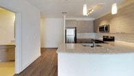 This well-appointed 2-bedroom, 2-bathroom single-floor residence offers a thoughtfully designed layout with modern finishes and a cohesive contemporary aesthetic. The open-concept living and kitchen area serves as the heart of the home, where rich hardwood-style flooring flows seamlessly underfoot. The kitchen is outfitted with sleek, textured flat-panel cabinetry in a warm taupe finish, a white subway tile backsplash, granite countertops, and a peninsula with an integrated sink — ideal for casual dining or entertaining. A full suite of stainless steel appliances, including a refrigerator, range, microwave, and dishwasher, completes the space. Pendant lighting and a track light fixture add both function and style overhead. The spacious living area is bathed in natural light from large sliding glass doors fitted with vertical blinds. Both bedrooms feature plush wall-to-wall carpeting, generous windows with blinds, and ample room for furnishings. The primary bedroom is accompanied by a full bathroom with a granite-topped vanity and a walk-in closet with built-in shelving. The second bathroom offers a tub-shower combination, a matching vanity, and large-format tile flooring. Clean white walls and neutral tones throughout create a bright, welcoming atmosphere.