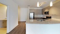 This well-appointed 2-bedroom, 2-bathroom single-floor residence offers a thoughtfully designed layout with modern finishes and a cohesive contemporary aesthetic. The open-concept living and kitchen area serves as the heart of the home, where rich hardwood-style flooring flows seamlessly underfoot. The kitchen is outfitted with sleek, textured flat-panel cabinetry in a warm taupe finish, a white subway tile backsplash, granite countertops, and a peninsula with an integrated sink — ideal for casual dining or entertaining. A full suite of stainless steel appliances, including a refrigerator, range, microwave, and dishwasher, completes the space. Pendant lighting and a track light fixture add both function and style overhead. The spacious living area is bathed in natural light from large sliding glass doors fitted with vertical blinds. Both bedrooms feature plush wall-to-wall carpeting, generous windows with blinds, and ample room for furnishings. The primary bedroom is accompanied by a full bathroom with a granite-topped vanity and a walk-in closet with built-in shelving. The second bathroom offers a tub-shower combination, a matching vanity, and large-format tile flooring. Clean white walls and neutral tones throughout create a bright, welcoming atmosphere.