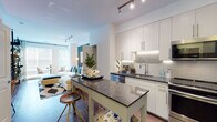This single-floor residence showcases a thoughtfully designed open-concept layout connecting the kitchen and living areas. The kitchen is a standout feature, equipped with stainless steel appliances including a French door refrigerator, gas range, built-in microwave, and dishwasher. Crisp white cabinetry extends to the ceiling, complemented by under-cabinet lighting, a grey subway tile backsplash, and dark granite countertops. A freestanding island with matching granite provides additional prep space and casual seating with bar stools. Rich wood-look flooring runs throughout the main living areas, adding warmth to the contemporary palette. Track lighting illuminates the kitchen and entryway, while the open living area benefits from natural light through large windows. The bedroom offers a dramatic contrast with a deep charcoal accent wall, plush carpeting, and a large window dressed with elegant curtains, creating a cozy and stylish retreat. The bathroom features large-format white wall tiles surrounding a soaking tub with shower, coordinating floor tile, warm wood-toned cabinetry, and a marble-look vanity surface, delivering a clean and functional design.