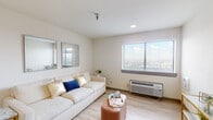 This single-floor unit features a thoughtfully designed layout that maximizes functionality within a compact footprint. The living area is bright and airy, with light wood-look flooring and crisp white walls that create a clean, neutral backdrop. A large window fitted with horizontal blinds allows natural light to fill the space while offering outdoor views, and a wall-mounted HVAC unit provides individual climate control. Two framed mirrors on the wall add depth to the room.

The bedroom continues the unit's consistent aesthetic, with the same light flooring and neutral walls. A window frames outdoor views, and the room comfortably accommodates a full-sized bed with coordinating nightstands. A glass-and-metal shelving unit provides additional storage.

The kitchen is efficiently designed with light wood-tone cabinetry, white subway tile backsplash, a smooth-top electric cooktop, a stainless steel sink, and a compact dishwasher. Open shelving above the counter adds visual interest and practical storage.

The bathroom features hexagonal tile flooring, a combined shower and tub with a curtain rod, a floating vanity with an integrated sink, and a framed mirror, completing the unit's cohesive, modern design.