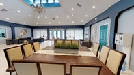This single-floor space makes a strong architectural statement with its vaulted ceilings that rise dramatically overhead, creating an airy, expansive atmosphere. A central skylight draws in abundant natural light, complementing the recessed lighting and an elegant chandelier that together illuminate the room warmly. The interior design is defined by bold navy blue accent walls contrasted against crisp white ceilings and light-toned flooring, giving the space a sophisticated coastal character. A striking teal-painted door adds a vibrant pop of color, reinforcing the space's distinctive palette. At the heart of the room sits a large, solid wood dining table surrounded by upholstered chairs with nailhead trim, centered with a decorative planter filled with ornamental grasses. The open floor plan flows seamlessly into a comfortable lounge area furnished with neutral-toned sofas and accent chairs, making the space equally suited for casual gatherings and formal dining. Along the far wall, a row of French doors and large windows dressed with white roller shades allow for generous natural light while maintaining privacy. Wall art and carefully placed accent pieces throughout add a refined, welcoming touch to this well-appointed communal space.