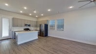 This two-story home showcases a contemporary design with clean lines and modern finishes throughout. The entry level features light wood-look flooring that extends through an open-concept layout, beginning at a bright foyer with a frosted-glass front door. The kitchen serves as a central highlight, equipped with gray shaker-style cabinetry, a white subway tile backsplash, stainless steel appliances including a gas range, microwave, and French door refrigerator, and a generously sized island topped with veined quartz countertops and an undermount sink. The open living area adjacent to the kitchen is bathed in natural light from multiple windows fitted with horizontal blinds. A staircase leads to the upper level, where the flooring transitions to plush carpet in the bedroom areas. Bedrooms are well-proportioned, each featuring ceiling fans, natural light from dual windows, and five-panel interior doors. The primary suite connects to a bathroom finished with marble-look tile flooring, a gray vanity with a white countertop, and a separate water closet. The overall aesthetic throughout is cohesive, blending neutral tones, modern hardware, and thoughtful finishes.
