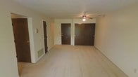 This single-floor residence offers a clean and functional layout designed for comfortable everyday living. The main room is finished with wall-to-wall carpeting in a neutral tone, creating a warm and cohesive atmosphere throughout. A ceiling fan with an integrated light fixture is centrally mounted, providing both air circulation and ambient lighting to the space.

Storage is a notable feature of this room, with a set of wide double-door closets along the back wall offering generous organizational capacity. An additional single door provides access to a secondary room or utility space, adding further versatility to the floor plan. The walls are painted in a soft, neutral palette that keeps the interior feeling light and open.

A floor-level ventilation register is visible along the baseboard, indicating a ducted heating and cooling system for year-round climate comfort. The textured ceiling adds a subtle architectural detail consistent throughout the space.

The overall ambiance is understated and move-in ready, with a practical design that emphasizes functionality. The neutral finishes and straightforward layout make this space highly adaptable to a variety of furnishing styles and personal preferences, appealing to those seeking a low-maintenance, well-organized living environment.
