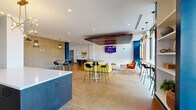 Located in Memphis, TN (38104), this single-floor clubhouse offers a well-appointed, open-concept layout designed for both socializing and relaxation. The space features polished concrete floors throughout, lending an industrial-chic aesthetic that pairs seamlessly with the modern interior design.

The lounge area is anchored by a bold yellow modular sectional sofa, a round upholstered ottoman, and a patterned area rug, all positioned beneath a striking wood-slat ceiling feature with integrated linear lighting and a wall-mounted flat-screen TV. Accent chairs in warm orange tones add a pop of contrast, completing a vibrant yet cohesive seating arrangement.

A dedicated dining zone features a long solid-wood communal table with pendant lighting overhead, ideal for group gatherings. Multiple high-top bar tables with sleek black stools are distributed throughout the space, offering flexible seating options. A kitchenette area includes a dark-finished island with a white countertop, a sink, and an additional wall-mounted TV.

Textured accent walls and bold cobalt blue columns punctuate the open floor plan, adding architectural depth. Floor-to-ceiling windows along one facade flood the space with natural light and provide views of the surrounding streetscape. Recessed lighting and designer pendant fixtures complete the polished, contemporary ambiance of this versatile clubhouse.