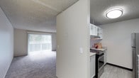 This single-floor residence offers a practical and comfortable layout designed for everyday living. The open-concept design connects the kitchen and living area, creating a sense of spaciousness throughout. The living area is finished with soft carpeting underfoot and benefits from abundant natural light streaming in through large sliding glass doors dressed with vertical blinds, which also provide access to an outdoor space. The textured ceiling adds a classic touch consistent throughout the home. The kitchen is efficiently arranged and features white upper cabinetry with open shelving, a stainless steel range with an overhead ventilation hood, and light-toned countertops that provide ample prep space. The transition between the kitchen and living area is defined by a partial dividing wall, lending a degree of separation while maintaining the open feel of the floor plan. Light-toned hard flooring in the kitchen contrasts with the carpeted living space, clearly delineating the two zones. Overall, the space presents a clean, neutral palette that provides a versatile backdrop for a variety of furnishing styles and personal touches.