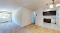 Located at 8 Baron Park Ln in Burlington, Massachusetts, this single-floor residential unit offers a functional and straightforward layout across several well-defined rooms. The open living area is finished with wall-to-wall carpeting and crisp white walls, creating a bright and neutral backdrop. A half-wall partition separates the living space from the kitchen, which features dark wood cabinetry extending to the upper walls, white countertops, a tiled backsplash, and white appliances including an electric range, dishwasher, and refrigerator. The kitchen floor is finished in a light-colored vinyl tile. The unit includes two carpeted bedrooms, each equipped with windows overlooking lush greenery and fitted with wall-mounted air conditioning units and baseboard heating. Both bedrooms offer generous closet space with louvered bi-fold doors. The full bathroom features classic tile wainscoting, a tiled tub/shower combination, and a small linen cabinet. A hallway connects the main living areas to the bedrooms and bathroom, with an electrical panel conveniently accessible from the corridor. Natural light filters throughout the unit via multiple windows framing views of the surrounding trees.