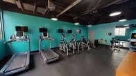 This fitness center is designed to provide a comprehensive workout environment within a single, open-plan floor. The space is immediately defined by its bold aesthetic: vibrant teal walls contrasted against a matte black exposed ceiling, which reveals structural ductwork, piping, and pendant-style overhead lighting. The dark rubber flooring throughout provides durability and traction suitable for high-intensity use.

The cardio zone features a row of treadmills and elliptical machines, each equipped with individual mounted flat-screen monitors, allowing users to stay entertained during their sessions. A utility station with a sink is conveniently positioned nearby. Adjacent to the cardio area, a dedicated strength training zone houses a multi-functional cable machine, weight benches, a leg press machine, and a power rack with barbell storage — catering to a full range of resistance training exercises.

Floor-to-ceiling mirrors line multiple walls of the strength area, visually expanding the space and allowing users to monitor their form. Decorative indoor plants add a touch of organic warmth to the otherwise industrial setting. Large windows and a glass entry door along one wall allow natural light to filter in, offering views of the surrounding outdoor environment. The overall ambiance is energetic and purpose-built, balancing functionality with a modern, urban fitness aesthetic.