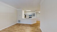 This single-floor residence features a clean, modern design characterized by light hardwood flooring, crisp white walls, and crown molding that runs throughout the main living areas. The open-concept layout connects the living area directly to the kitchen, which is defined by a peninsula counter with a built-in sink, offering both additional workspace and a natural division between the two areas. The kitchen is equipped with stainless steel appliances, including a refrigerator, range, and over-the-range microwave, complemented by gray cabinetry with brushed nickel hardware and white countertops. Ample upper and lower cabinet storage lines the kitchen walls, with a separate interior door providing additional access or storage.

The living area is generously proportioned, allowing flexible furniture arrangements, while the entry area benefits from a large window that invites natural light and outdoor views. The bedroom is well-sized, featuring a closet and a window overlooking what appears to be an exterior balcony or patio area. The bathroom includes a full soaking tub with white tile surround, a toilet, and a vanity area. Consistent hardwood-style flooring flows throughout, contributing to a cohesive and bright overall ambiance.