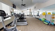 This fitness center is housed in a single-story space designed to support a comprehensive workout experience. The room features soaring ceilings with clerestory-style windows along the upper walls, allowing abundant natural light to fill the space and creating an open, airy atmosphere. Two ceiling fans are installed overhead, promoting air circulation throughout the room.

The cardio section is prominently lined with a row of Cybex treadmills and elliptical machines positioned along one side of the room. A wall-mounted flat-screen television is conveniently placed for viewing during workouts. Toward the back of the room, a free weights and strength training area features dumbbells, weight benches, and additional resistance equipment. A large floor-to-ceiling mirror runs along one wall, visually expanding the space while allowing users to monitor their form during exercise.

The flooring consists of patterned carpeting suited for a fitness environment. Exercise accessories such as stability balls and step platforms are also available. A wall clock is mounted for easy time management. The overall ambiance is clean, functional, and well-lit, making it suitable for a wide range of fitness activities.