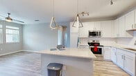 This single-floor residence showcases a thoughtfully designed open-concept layout with consistent light wood-style flooring connecting the main living areas. The kitchen is a standout feature, outfitted with crisp white shaker cabinetry extending to the ceiling, a classic white subway tile backsplash, and a full suite of stainless steel appliances including a range, microwave, dishwasher, and refrigerator. A spacious central island with a light quartz countertop provides additional workspace and casual seating. Two geometric triangular pendant lights in a brushed gold finish hang above the island, adding a contemporary focal point, while track lighting further illuminates the space.

The living area features neutral gray walls and light-toned flooring, offering a clean and versatile backdrop. Bedrooms are carpeted and receive natural light through double-pane windows, with outdoor views of surrounding greenery. A dedicated laundry room houses a full-size washer and dryer with built-in shelving above. A glazed exterior door in the living area allows additional natural light and outdoor access. Throughout the space, white trim, neutral tones, and modern gold-accented fixtures create a cohesive, contemporary aesthetic.