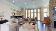 This elegantly designed single-floor space showcases a seamless open-concept layout that connects the kitchen, dining, and living areas in one cohesive, flowing environment. The soaring ceilings amplify the sense of grandeur, while floor-to-ceiling windows dressed in sheer gold drapery bathe the interior in generous natural light and offer views of the surrounding exterior. The gourmet kitchen is outfitted with sleek cabinetry, a dark tile backsplash, stainless steel appliances, and a spacious island with bar seating — all illuminated by a trio of stylish pendant lights. Dark hardwood flooring runs throughout, providing a rich contrast to the light-toned walls and furnishings. The living area is anchored by a large neutral sectional sofa accented with warm orange and patterned throw pillows, centered around a wood coffee table atop a traditional area rug. A dedicated dining area near the windows accommodates comfortable seating for multiple guests. Recessed lighting supplements the natural light, ensuring the space remains warm and inviting throughout the day. The overall ambiance is refined yet welcoming, blending classic architectural details with contemporary finishes.