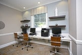 This single-floor space is designed to serve as a functional yet stylish shared workspace and lounge area. The room is finished with neutral gray walls, white wainscoting, and crown molding that lend a polished, professional atmosphere throughout. Recessed lighting is installed across the ceiling, providing consistent, even illumination across the entire space.

Along one wall, a built-in desk runs beneath a centered window dressed with a simple valance, allowing natural light to filter into the room. Three workstations are arranged along this desk, each equipped with a desktop computer and accompanied by modern ergonomic chairs with wood accents. Floating shelves flank the window on either side, displaying minimal decorative accessories that contribute to the clean, contemporary aesthetic.

To the right, a high-top round glass table with white bar-height chairs offers an informal seating area suited for casual meetings or collaborative work. The flooring throughout features a light-toned tile in a parquet-style pattern, complementing the overall neutral palette. The space balances practicality with refined design, making it well-suited for productivity and informal gatherings alike.