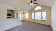 This single-story home showcases an open-concept living and dining area defined by a dramatic vaulted ceiling that creates a strong sense of spaciousness and volume. A ceiling fan with integrated lighting is centrally positioned overhead, while a distinctive arched transom window near the peak of the vaulted ceiling serves as a focal architectural detail, drawing in natural light from above. Multiple large windows along the exterior walls further enhance the brightness of the space, offering views of the surrounding greenery outside.

The living area features wall-to-wall carpeting, providing a warm and comfortable foundation underfoot, while the dining area transitions to hard surface flooring, clearly delineating the two zones within the open layout. A pass-through counter connects the living and dining spaces to the kitchen, which is partially visible and features cabinetry in a classic style. A decorative chandelier is positioned above the dining area, adding a refined touch to the open floor plan.

The walls are finished in a soft neutral tone, complementing the textured ceiling treatment throughout. The overall ambiance of the space is bright, airy, and welcoming, with natural light playing a central role in the character of the interior.