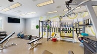 Located at 2 Abbott Dr in Hampton, Virginia, this fitness center offers a well-appointed exercise space spread across a single floor. The facility is divided into distinct workout zones, each designed to support a comprehensive fitness routine.

The cardio area is bright and airy, lined with large white-framed windows fitted with plantation shutters that allow abundant natural light and views of the surrounding greenery. This section houses multiple treadmills and elliptical machines, complemented by air resistance bikes. Ceiling fans overhead and wall-mounted flat-screen televisions enhance the comfort of cardio workouts.

A separate strength and free weights area features a full dumbbell rack, an adjustable workout bench, and a cable/multi-use strength machine. A wall-mounted flat-screen TV and additional natural light from French doors leading to the outdoors make this zone both functional and inviting.

The strength training zone is highlighted by a bold, floor-to-ceiling motivational mural that energizes the space. This area includes a power rack with a barbell and weight plates, adjustable benches, and additional strength equipment, all set on durable speckled flooring throughout the facility.

Recessed lighting fixtures and ceiling fans are distributed across the entire space, ensuring consistent illumination and airflow. The clean, light-toned walls in soft blue and white tones create a fresh, motivating atmosphere suited for a wide range of fitness activities.