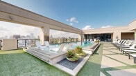 Located at 2511 Willowick Rd in Houston's upscale 77027 corridor, this rooftop amenity deck offers a compelling blend of resort-style leisure and urban sophistication. The centerpiece is a sleek, elongated pool set against a backdrop of the Houston skyline, visible in stunning panoramic fashion from nearly every vantage point on the deck. Surrounding the pool, a generously sized sun terrace is finished with a combination of stone pavers and synthetic turf, lined with an array of contemporary lounge chairs designed for relaxed sunbathing. Lush potted planters and manicured greenery add refined touches of color throughout the space. A covered outdoor lounge area features a warm wood-paneled ceiling, large ceiling fans for comfort, and plush sectional seating — ideal for social gatherings or quiet relaxation. Adjacent to the lounge, a fully equipped outdoor kitchen includes a stainless steel grill, prep station, and a mounted flat-screen television, complemented by a dining table with stylish woven chairs. The architectural detailing throughout — including exposed brick columns, floor-to-ceiling glass partitions, and clean modern lines — reinforces the elevated character of this outdoor amenity space.