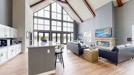 Located at 700 S Main St in Oshkosh, Wisconsin, this well-appointed single-floor community space offers a generous open layout designed for both gathering and relaxation. The architecture is immediately striking, with dramatically vaulted ceilings accented by rich, exposed wood beams that lend a warm yet contemporary character throughout the main living area.

The combined kitchen and living room serves as the heart of the space. The kitchen features white shaker cabinetry, granite countertops, stainless steel appliances, and a large island with bar seating — ideal for casual dining or entertaining. The adjacent living area centers around a stacked-stone fireplace with a wood mantel, flanked by comfortable seating and bathed in natural light from multiple windows.

A dedicated dining room provides flexible high-top table seating with warm leather swivel chairs, complemented by a billiards table and wall-mounted TV — creating a dynamic lounge atmosphere. A sliding barn door adds a stylish architectural detail to this area.

At the far end of the space, dramatic floor-to-ceiling glass curtain walls and French doors open directly to an outdoor pool area, flooding the interior with light and providing seamless indoor-outdoor connectivity. Light wood-look flooring runs throughout, tying each area together cohesively. The overall ambiance is modern, airy, and inviting.