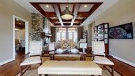 This single-story clubhouse is designed to deliver both sophistication and functionality across a range of thoughtfully appointed spaces. The grand main lounge serves as the centerpiece, showcasing a striking coffered ceiling with rich dark wood beams, recessed lighting, and an oversized industrial pendant light. A dramatic floor-to-ceiling stacked stone fireplace anchors one wall, while large French doors flood the room with natural light and offer views to the outdoors. Plush seating arrangements rest atop natural fiber rugs over hardwood floors, creating an inviting atmosphere. An adjacent lounge area features neutral-toned furnishings, floor-length drapery, and windows overlooking a pool area. A formal dining nook accommodates a round table with upholstered chairs and botanical wall art. The kitchen is outfitted with granite countertops, white cabinetry, stainless steel appliances, and a bar-height island. The fitness center includes a variety of strength machines set against large windows, while a separate studio room provides light wood flooring, cycling bikes, and wall-mounted exercise balls for group fitness activities. An additional multipurpose room with a projection screen rounds out the facility.