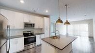This well-appointed 2-bedroom, 2-bathroom residence combines modern design with functional living across a single floor. The open-concept kitchen serves as the heart of the home, featuring crisp white cabinetry reaching to the ceiling, dark quartz countertops, a hexagonal tile backsplash, stainless steel appliances, and a central island with a built-in sink topped by a stylish pull-down faucet. Two brushed gold dome pendant lights hang above the island, adding a warm, contemporary accent. The kitchen flows naturally into the living area, which is finished with wood-look flooring and a large sliding glass door fitted with vertical blinds that invite natural light. Both bedrooms are carpeted for comfort, with the primary bedroom featuring a distinctive sliding barn door that opens to a dedicated closet and en-suite bathroom. The primary bath includes a white vanity with dark countertops, an undermount sink, and hardwood-style flooring. A second carpeted bedroom and additional full bathroom offer flexibility for guests or shared living. Two closets provide ample storage, and an in-unit washer and dryer are included. The overall aesthetic is clean, neutral, and modern throughout.