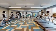 Located at 3575 Silverside Rd in Wilmington, Delaware, this well-appointed two-floor fitness center offers a comprehensive workout environment designed for both performance and comfort. The space is defined by its modern aesthetic, featuring recessed linear LED lighting across smooth white ceilings and large windows that flood the interior with natural light while providing views of the surrounding area.

The main cardio floor is expansive, accommodating a full array of equipment including treadmills, ellipticals, stair climbers, and stationary bikes. A striking abstract mural adorns one wall, adding an artistic focal point to the space. The floors are covered in a bold, geometric-patterned carpet in blue, tan, and grey tones that lends energy and visual interest to the environment. A full-length mirror along one wall further enhances the sense of openness.

A dedicated strength and selectorized machine area features rubberized flooring and a wide selection of resistance equipment, all arranged to maximize usability and circulation. This zone is equally well-lit, with large windows offering outdoor views. A functional training corner provides stability balls, resistance tools, and other accessories.

The second floor is accessible via a staircase and extends the facility's footprint. Throughout both levels, the design maintains a cohesive, contemporary style with neutral grey walls, wood-tone base trim, and thoughtfully organized equipment layouts suited for a broad range of fitness activities.