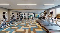 Located at 3575 Silverside Rd in Wilmington, Delaware, this well-appointed two-floor fitness center offers a comprehensive workout environment designed for both performance and comfort. The space is defined by its modern aesthetic, featuring recessed linear LED lighting across smooth white ceilings and large windows that flood the interior with natural light while providing views of the surrounding area.

The main cardio floor is expansive, accommodating a full array of equipment including treadmills, ellipticals, stair climbers, and stationary bikes. A striking abstract mural adorns one wall, adding an artistic focal point to the space. The floors are covered in a bold, geometric-patterned carpet in blue, tan, and grey tones that lends energy and visual interest to the environment. A full-length mirror along one wall further enhances the sense of openness.

A dedicated strength and selectorized machine area features rubberized flooring and a wide selection of resistance equipment, all arranged to maximize usability and circulation. This zone is equally well-lit, with large windows offering outdoor views. A functional training corner provides stability balls, resistance tools, and other accessories.

The second floor is accessible via a staircase and extends the facility's footprint. Throughout both levels, the design maintains a cohesive, contemporary style with neutral grey walls, wood-tone base trim, and thoughtfully organized equipment layouts suited for a broad range of fitness activities.