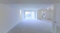 This single-floor residence is designed with an open, airy layout that maximizes the sense of space and light throughout. The main living area is expansive and finished with plush wall-to-wall carpeting, creating a comfortable and welcoming atmosphere. Crisp white walls reflect natural light generously, contributing to the overall brightness of the interior.

A defining feature of the living area is the large sliding glass door dressed with vertical blinds, which floods the space with abundant natural light and provides direct access to an outdoor area. The view through the glass offers a pleasant outdoor backdrop, enhancing the connection between the interior and exterior.

Seamlessly accessible from the main living space, the kitchen is efficiently laid out and equipped with classic white appliances, including a range and microwave, set against warm wood-toned cabinetry. The neutral flooring in the kitchen complements the overall light color palette of the home.

The open flow between the living and kitchen areas makes this space well-suited for everyday living and easy entertaining. The clean, neutral finishes throughout provide a versatile canvas, allowing for a wide range of interior design possibilities. Overall, the space presents a bright, functional, and inviting environment.