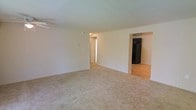 This single-floor space features an open and functional layout designed for comfortable everyday living. The main living area is finished with wall-to-wall neutral-toned carpeting, providing a warm and understated foundation. Smooth, light-colored walls and a standard ceiling with a ceiling fan contribute to a clean, simple aesthetic throughout the room.

Toward the back of the living area, an open passthrough connects directly to the kitchen, which transitions to hardwood-style flooring. The kitchen is equipped with a full-size black refrigerator, visible from the living space, offering a practical and accessible layout. The open connection between the two areas promotes a natural flow, making the overall floor plan feel cohesive.

A hallway accessible from the living area leads further into the unit, suggesting additional rooms beyond what is immediately visible. The space is well-lit with neutral ambient lighting, and the overall color palette of whites and beiges lends the interior a clean, move-in-ready appearance. The layout is straightforward and efficient, suited for a range of furnishing arrangements.