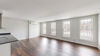 Located at 244 Worthington St in downtown Springfield, MA, this one-bedroom, one-bathroom unit offers a well-designed single-floor layout with a clean, modern aesthetic. Rich, dark hardwood flooring runs consistently throughout the space, creating a cohesive and polished look.

The open-concept kitchen and living area forms the heart of the unit. The kitchen is equipped with white shaker cabinetry, dark granite countertops, stainless steel appliances including a refrigerator and range, and a striking island with a pendant range hood — ideal for both cooking and entertaining. The living area is generously proportioned, bathed in natural light from a row of large double-hung windows that offer views of the surrounding urban streetscape.

The bedroom is a comfortable, well-lit space featuring its own ductless mini-split unit for individualized climate control, along with a window that brings in natural light. Ductless mini-split systems are present throughout the unit, ensuring efficient heating and cooling in every room.

The bathroom features a full tub-shower combination, warm earth-toned tile wainscoting, and tile flooring, giving the space a clean and functional finish. The overall design is neutral and modern, with white walls providing a bright, inviting atmosphere throughout.
