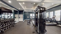 Located at 10880 Angel Fish Way in Jacksonville, FL, this fitness center offers a comprehensive workout environment designed with both functionality and aesthetics in mind. The space is defined by its striking deep teal-blue walls, crisp white crown molding, and coffered ceilings with recessed lighting, creating a polished and motivating atmosphere throughout.

The main workout floor is expansive, finished with durable rubber flooring and flooded with natural light from multiple large windows fitted with blinds. A row of modern treadmills with individual screens lines one wall, complemented by additional cardio equipment including ellipticals and stationary cycles. A Matrix multi-station functional trainer serves as a centerpiece, surrounded by adjustable weight benches and a variety of strength machines for a full-body workout.

A dedicated free weights area features a full rack of dumbbells displayed in front of a large wall mirror, ideal for both form monitoring and space amplification. Built-in white cabinetry and open shelving provide convenient storage. A separate secondary area accommodates spin bikes and additional benches, with signage, an AED unit, and a fire extinguisher for safety compliance.

A well-lit hallway connects ancillary rooms, maintaining the same clean, neutral tile flooring and consistent design language. Ceiling fans throughout ensure comfortable air circulation, rounding out this thoughtfully equipped and attractively appointed fitness facility.