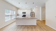 Located at 5895 Arbor Isle Way in Myrtle Beach, South Carolina, this single-floor residence offers a clean, modern design with a bright and airy atmosphere. The open-concept layout seamlessly connects the kitchen and living areas, creating a spacious, functional flow throughout the home. Light wood-look flooring runs consistently across the entire space, enhancing the sense of continuity and warmth.

The kitchen is a standout feature, anchored by a large center island with a granite countertop and an undermount sink, illuminated by a row of pendant lights with gold-toned fixtures. White shaker-style cabinetry lines the walls, complemented by a subway tile backsplash and stainless steel appliances, including a refrigerator, range, and microwave. A built-in wine rack adds a refined touch above the refrigerator.

The living area is generously proportioned, featuring a ceiling fan and access to the outdoors through French doors, with additional windows allowing natural light to pour in. The bedroom continues the light, neutral palette with its own ceiling fan and a large window overlooking the surroundings. A spacious walk-in closet with wire shelving provides excellent storage capacity. The bathroom is finished with a granite-topped vanity, wood-look flooring, and a full-width glass-door walk-in shower with tiled walls and built-in corner shelving.