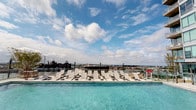 This rooftop amenity floor presents a thoughtfully designed communal environment that blends open-air leisure with polished interior spaces. The centerpiece is a generously sized rooftop pool with crystal-clear turquoise water, framed by a wide pool deck lined with rows of contemporary lounge chairs. The elevated position affords expansive, unobstructed views of the surrounding urban skyline, creating a resort-like atmosphere above the city.

Adjacent to the pool, a dedicated outdoor lounge area features modular seating arrangements and a central coffee table set atop large-format stone pavers. Lush planters and raised greenery add a natural element to the terrace, enhancing its ambiance. The space operates during set daytime hours and includes dedicated pool equipment access rooms, indicating well-maintained infrastructure behind the scenes.

The interior corridor leading to the rooftop amenities is equally refined, featuring light-toned large-format tile flooring, crisp white walls, and recessed lighting. Framed artwork lines the hallway, introducing a gallery-like quality to the transitional space. Access-controlled entry points throughout reflect a secure and professionally managed environment. The overall aesthetic is clean, modern, and cohesive.