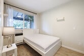 Located at 1831-1835 Harmon St in Berkeley, California, this single-room space is a thoughtfully designed bedroom that maximizes every inch of its compact footprint. The room features light gray-beige walls and warm-toned laminate wood flooring, creating a clean, neutral aesthetic that feels bright and inviting. A sliding-panel wardrobe with frosted glass doors provides practical built-in storage, while a wall-mounted shelf with gold bracket hardware adds a functional decorative touch. Natural light enters through a single window dressed with sheer white curtains and a black curtain rod, offering a pleasant view of the lush, tree-lined outdoor area — a covered porch with visible exposed beams and greenery. A wall-mounted heater unit sits beneath the window, ensuring climate comfort year-round. The room also includes a slide-in entry door, adding privacy and space efficiency. The overall ambiance is minimal and modern, with a calm, neutral palette that lends itself well to a restful environment. This self-contained bedroom is well-suited for single occupancy and reflects a practical, clean design sensibility fitting for the Berkeley area.
