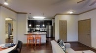 This single-story space showcases an open-concept design that seamlessly connects the kitchen and living area, creating a cohesive and functional layout. The kitchen is outfitted with dark espresso cabinetry that extends to the upper walls, providing ample storage. Stainless steel appliances, including a refrigerator, range, and microwave, add a modern touch. The kitchen features a peninsula with a granite-style countertop, illuminated by pendant lighting that hangs above, offering casual seating for two with vibrant orange bar stools that add a pop of color. A stainless steel sink is positioned along the peninsula, and a tile backsplash completes the kitchen's polished look. Recessed and flush-mount ceiling lighting ensures the space is well-lit throughout. Rich, dark hardwood-style flooring spans the entire area, grounding the design with warmth and continuity. Crown molding lines the ceiling, adding an architectural detail that elevates the overall aesthetic. The adjoining living area is spacious enough to accommodate comfortable seating and is accented by a neutral-toned sofa. A paneled door provides access to an additional room, completing this well-appointed space.