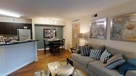 This single-floor residence offers a thoughtfully designed open-concept layout that seamlessly connects the living, dining, and kitchen areas into one cohesive living space. The living area is anchored by a plush gray sofa adorned with patterned accent pillows, complemented by a glass-top coffee table and warm ambient lighting. Framed wall art adds a polished finishing touch to the neutral-toned walls.

The dining area is defined by a rich accent wall in a deep sage green, creating a striking focal point within the open floor plan. A dark wood dining table with matching high-back chairs sits beneath a sleek track lighting fixture, providing both functionality and style. A decorative mirror and floral arrangement enhance the elegance of the dining nook.

Rich hardwood flooring flows throughout the main living areas, adding warmth and continuity to the space. The kitchen, partially visible behind a breakfast bar with dark countertops, integrates smoothly into the open plan. Ceiling-mounted HVAC vents and recessed lighting ensure year-round comfort. Overall, the space presents a modern, inviting atmosphere with a harmonious blend of warm tones, contemporary furnishings, and clean architectural lines.