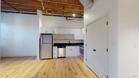 Located at 87 Washington St in Haverhill, MA, this thoughtfully renovated unit masterfully blends industrial character with contemporary design. The open living area makes a striking impression with soaring exposed timber beam ceilings, visible ductwork, and warm natural light filtering through large black-framed windows set against original exposed brick walls. Light-toned hardwood flooring flows seamlessly throughout, enhancing the sense of openness.

The kitchen is well-appointed with white shaker-style cabinetry, dark countertops, a hexagonal tile backsplash, a natural wood floating shelf, and a full suite of stainless steel appliances including a refrigerator, dishwasher, range, and microwave. Track lighting overhead provides both task and ambient illumination.

The bedroom continues the industrial-loft aesthetic with exposed brick accent walls and black-framed windows that invite abundant natural light. The bathroom offers a clean, modern finish with patterned geometric tile flooring, a floating wood-toned vanity with an integrated sink, a large mirror, and white subway tile surrounding the tub and shower. In-unit laundry is also accessible nearby.

This single-floor unit delivers a distinctive urban loft experience, combining historic architectural elements with high-quality modern upgrades in a compelling Haverhill location.