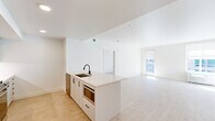 Located in Portland's vibrant NW District, this two-bedroom, two-bathroom residence offers a clean, modern aesthetic across a well-designed single-floor layout. The open-concept kitchen and living area serves as the heart of the home, anchored by a sleek white kitchen island with a black matte faucet and integrated sink. The kitchen is well-appointed with white shaker-style cabinetry, a tiled backsplash, stainless steel appliances including a full-size refrigerator and gas range, and recessed lighting throughout. Light wood-tone flooring extends seamlessly across all rooms, enhancing the bright and airy feel. The living area receives generous natural light through large windows and a glass door, with views of the surrounding urban environment. The primary bedroom is a comfortable retreat, featuring multiple windows, a dedicated bathroom with white cabinetry, quartz countertops, black hardware accents, and access to a walk-in closet with built-in shelving and hanging rods. The second bedroom is equally bright, with a well-positioned window that invites natural light. A second full bathroom serves the additional bedroom. The overall design is characterized by a crisp white palette, contemporary fixtures, and a cohesive, minimalist sensibility throughout.