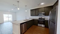 This thoughtfully designed one-bedroom, one-bathroom unit offers an efficient and functional layout across a single floor. The open-concept kitchen and living area forms the heart of the space, creating a seamless flow ideal for everyday living. The kitchen is outfitted with dark espresso cabinetry, granite countertops, a peninsula with an undermount sink, and a full suite of stainless steel appliances including a refrigerator, electric range, built-in microwave, and dishwasher. Pendant lighting above the peninsula adds a modern touch, while warm hardwood-style flooring runs throughout the entire unit, providing visual continuity and easy maintenance. The living area is open and airy, bathed in natural light, and features sliding glass doors that lead to a private balcony overlooking the surrounding area. The spacious primary bedroom includes a ceiling fan, a window with blinds, and ample room for a full furniture arrangement. The bathroom is well-appointed with a granite-topped dark vanity, a tiled walk-in shower, and a walk-in closet accessible directly from the bathroom. Clean white walls and recessed lighting throughout contribute to a bright, contemporary ambiance.