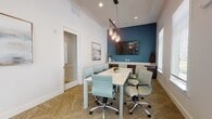 This single-floor professional space is thoughtfully designed to balance functionality with contemporary style. The layout includes a dedicated conference room and an open reception or lobby area, each with distinct finishes that contribute to a cohesive, polished aesthetic.

The conference room is a standout feature, centered around a sleek white rectangular table surrounded by cushioned light blue ergonomic chairs with chrome bases. A bold teal accent wall serves as a focal point, anchoring a wall-mounted flat-screen display ideal for presentations. A rustic wood-and-mason-jar pendant light fixture hangs overhead, adding warmth and character to the otherwise clean, modern design. Recessed lighting supplements the ambiance, while a built-in cabinet with a light countertop along the teal wall provides practical storage and display surface. Herringbone-patterned carpet throughout the conference area adds texture underfoot. Framed abstract artwork adorns the white walls, and a window with horizontal blinds introduces natural light.

The adjacent reception area contrasts with light tile flooring and soaring ceilings that amplify the sense of openness. Floor-to-ceiling glass walls and a glass entry door flood the space with natural light, offering views to an outdoor area. The transition between spaces is defined by a glass partition door, maintaining visual connectivity while delineating each zone clearly.