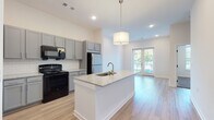 This single-floor residence features a thoughtfully designed open-concept layout where the kitchen and living area flow seamlessly together. The kitchen is equipped with gray shaker-style cabinetry offering generous storage, granite-look countertops, a white subway tile backsplash, and black appliances including a range, over-the-range microwave, and stainless steel refrigerator. A central island with a built-in sink and pendant lighting serves as both a functional workspace and a visual focal point. Light wood-look flooring extends throughout the main living areas, complementing the bright, neutral color palette and recessed lighting overhead. The living area benefits from notable ceiling height, enhancing the sense of openness, and is anchored by French doors that provide access to a private balcony with outdoor views. The bedroom features plush carpet flooring and a large window that brings in natural light. A well-appointed bathroom includes a granite-topped vanity, wood-look flooring, and a full soaking tub with shower. A walk-in closet with wire shelving provides practical storage, and an in-unit washer is conveniently located nearby.
