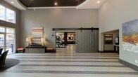 This single-floor space makes a strong first impression with its expansive, open layout and soaring ceilings that lend an airy, upscale feel. The interior design blends modern and industrial elements seamlessly, highlighted by a large, quilted sliding barn door mounted on a bold black metal track — a standout architectural feature that serves as both a functional divider and a focal point of the space. The flooring features wide, horizontal striped patterning in neutral tones, adding visual depth and a sense of movement throughout the area. Walls are finished in a soft gray palette, creating a clean and sophisticated backdrop. Recessed lighting is embedded in the dark-toned ceiling, providing focused illumination that enhances the room's ambiance. A tasteful console table, decorative lamp, and upholstered stool offer welcoming accents near the entrance. Beyond the barn door, a secondary lounge or gathering area is visible, featuring exposed brick-style walls, pendant lighting, and casual seating, suggesting a warm and inviting atmosphere. A colorful abstract artwork adds a vibrant contrast against the neutral tones, reinforcing the space's contemporary character. Overall, the design balances openness with defined zones, making it well-suited for a communal or commercial setting.