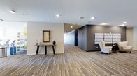 This well-appointed single-floor space presents a clean, contemporary aesthetic that blends comfort and functionality. The main corridor and lounge area are anchored by patterned carpet flooring in warm neutral tones, lending a sense of texture and visual interest throughout. Recessed lighting is installed throughout the ceiling, providing consistent, even illumination across the space.

A decorative console table along one wall is styled with an ornate framed mirror, a dark ceramic vase, and candle holders, adding a refined, curated touch to the entryway or lounge area. Adjacent to this, an accent wall in deep charcoal grey creates a bold visual contrast against the lighter surrounding walls, defining different zones within the open layout.

Comfortable seating with patterned upholstery is positioned near the accent wall, offering a relaxed area for occupants to gather. The corridor extends toward the back of the space, hinting at additional rooms or amenities beyond. Overall, the space strikes a balance between modern design sensibility and functional layout, with thoughtful décor choices, a neutral color palette, and architectural elements that contribute to a polished and inviting atmosphere.