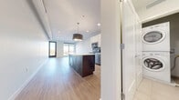 This contemporary single-floor residence offers a thoughtfully designed open-concept layout that seamlessly connects the kitchen and living areas. The kitchen is a standout feature, anchored by a substantial dark-cabinetry island with a crisp white countertop and a stylish pendant light overhead. Stainless steel appliances, including a microwave and range, are complemented by white upper cabinetry, creating a clean, modern aesthetic. Light wood-toned flooring flows throughout the main living spaces, enhancing the bright and airy atmosphere. Large floor-to-ceiling windows and a glass balcony door flood the living area with natural light while offering access to a private balcony overlooking an expansive urban cityscape. The bedrooms are carpeted for comfort, with one featuring a generous walk-in closet equipped with wire shelving and hanging rods. The bathroom includes a dark-cabinetry vanity with a white countertop, a soaking tub, and patterned tile surround. A dedicated vanity area with additional dark cabinetry and a large mirror provides further convenience. Throughout the unit, neutral tones, clean lines, and recessed lighting contribute to a polished, move-in-ready environment.