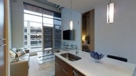 This contemporary single-floor residence showcases a thoughtfully designed open-concept layout that seamlessly integrates the kitchen, living, and sleeping areas. The kitchen is anchored by a clean-lined island with a white countertop and undermount stainless steel sink, complemented by warm wood cabinetry and modern pendant lighting that adds an elegant focal point overhead. The open living space benefits from dramatic floor-to-ceiling windows that flood the interior with natural light and offer expansive urban views, creating a strong sense of openness and connection to the city beyond. Polished concrete flooring runs throughout, lending an industrial-chic aesthetic that ties the space together. The living area features a wall-mounted flat-screen television and glass accent furniture that keep the space feeling light and uncluttered. The sleeping area, partially visible through an open passage, offers a warm, intimate retreat with soft ambient lighting and ample closet storage. The overall ambiance is clean, modern, and urban — a well-appointed space that balances functionality with contemporary style.