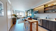 This single-floor residence showcases a thoughtfully designed open-concept layout with a seamless flow between the kitchen, dining, and living areas. The kitchen is outfitted with stainless steel appliances, two-tone wood cabinetry with under-cabinet LED strip lighting, white subway tile backsplash, and a central island/dining table with bar seating — ideal for casual dining and entertaining. Light wood-style flooring runs throughout, lending a warm, cohesive aesthetic.

The living area is anchored by a bold deep-green accent wall adorned with a curated gallery of framed artwork in gold frames, creating a striking focal point. Large windows along the exterior wall flood the space with natural light while offering urban cityscape views, enhancing the sense of openness.

The bedroom features the same muted green accent wall and large floor-to-ceiling windows with city views, creating a calm and airy retreat. A walk-in closet provides generous storage. The bathroom includes a tub-shower combination with white subway tile surround, light wood-look flooring, and clean, understated finishes throughout. The overall design balances contemporary style with warm, livable character.