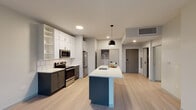 This single-floor space showcases a contemporary design with a cohesive neutral palette, light wood-tone flooring, and clean architectural lines throughout. The open-concept kitchen is a central highlight, featuring two-tone cabinetry — white upper cabinets with dark lower cabinets — complemented by a white subway tile backsplash, stainless steel appliances including a French door refrigerator, electric range, and built-in microwave. A substantial kitchen island with a crisp white countertop and a deep navy base provides additional workspace and includes an integrated sink, pendant lighting above adds a modern touch. Adjacent to the kitchen, a dedicated utility area houses a stacked washer and dryer unit alongside open shelving, accessed through a stylish sliding barn door. The bathroom is well-appointed with a dark-toned vanity, white countertop, a large mirror, recessed wall shelving, and a glass-enclosed shower. The living areas are bright and airy, with large windows offering street-level urban views, while a door leads to an outdoor balcony. Additional closet storage is thoughtfully integrated near the entry.